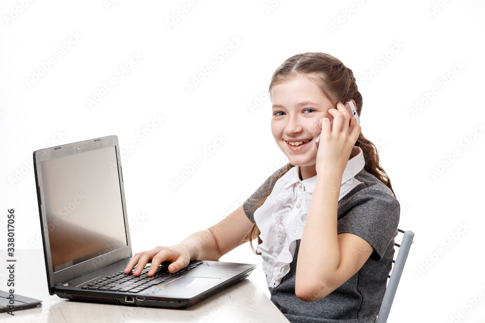 Cute schoolgirl sitting at a laptop and talking on the phone on a white background Cute schoolgirl sitting at a laptop and talking on the phone on a white background