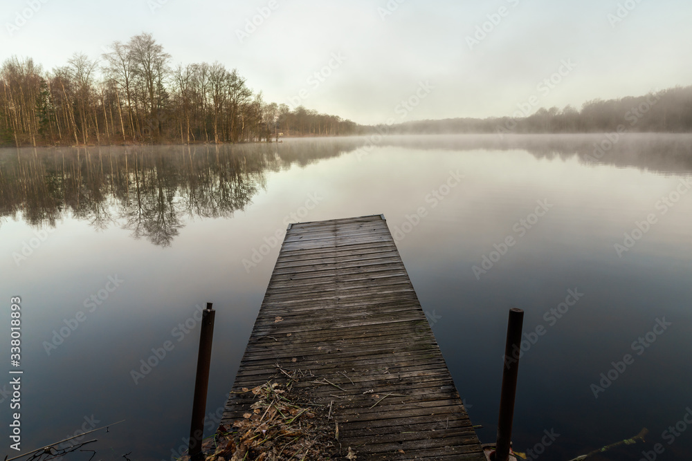 Naklejka premium A jetty by a mirrored lake on an early morning with fog easing