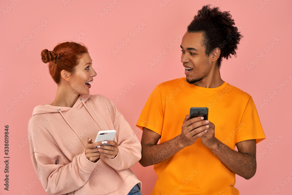 Image of couple African American man and ginger caucasian woman smiling while both using mobile phones isolated over pink background. Modern technology, communication, and relationships. Image of couple African American man and ginger caucasian woman smiling while both using mobile phones isolated over pink background. Modern technology, communication, and relationships.