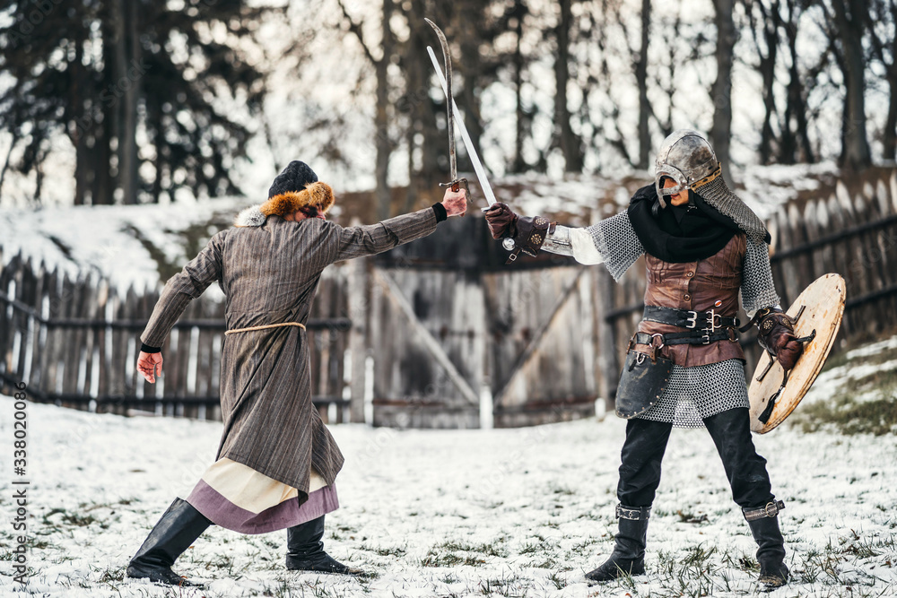 Fototapeta premium Two ancient warriors in armor with weapons fighting with swords in the snow
