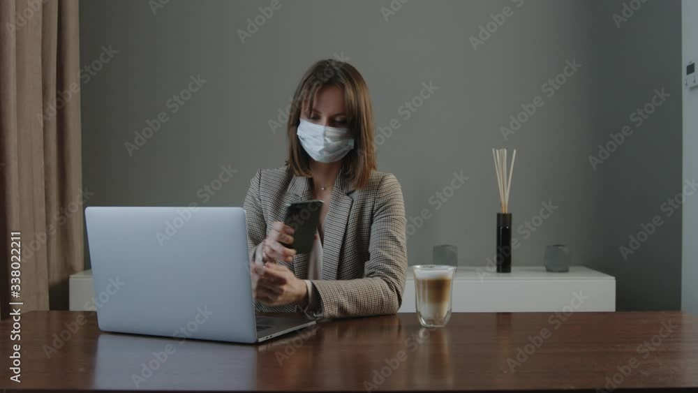 Young Woman in a mask and jacket works from home on a laptop at a table and talks on the phone in quarantine Young Woman in a mask and jacket works from home on a laptop at a table and talks on the phone in quarantine