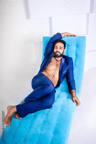 Attractive happy Asian young bearded hairy shirtless male model wearing dark-blue suit having rest on light-blue coach or sofa © arrideo