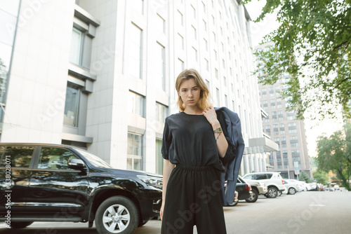 Outdoor fashion portrait of young businesswoman posing in the street of european city near the car. Stylish businesswoman standing in front of the business center holding her jacket.