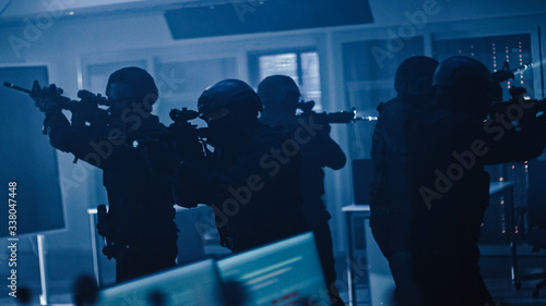 Masked Squad of Armed SWAT Police Officers Stand in Dark Seized Office Building with Desks and Computers. Soldiers with Rifles and Flashlights Surveil and Cover Surroundings. © Gorodenkoff
