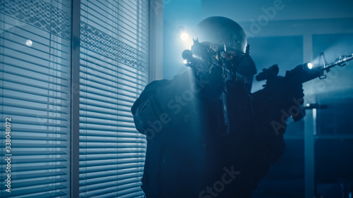 Close Up Portrait of Masked Armed SWAT Police Officer Storm a Dark Seized Office Building with Desks and Computers. Soldiers with Rifles and Flashlights Move Forward and Cover Surroundings.