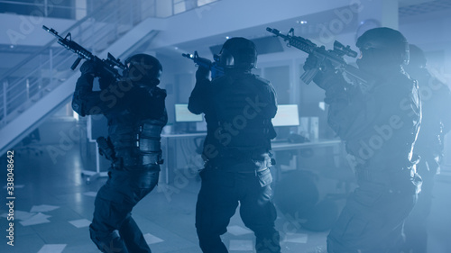 Masked Team of Armed SWAT Police Officers Slowly Move in a Hall of a Dark Seized Office Building with Desks and Computers. Soldiers with Rifles and Flashlights Surveil and Cover Surroundings. © Gorodenkoff