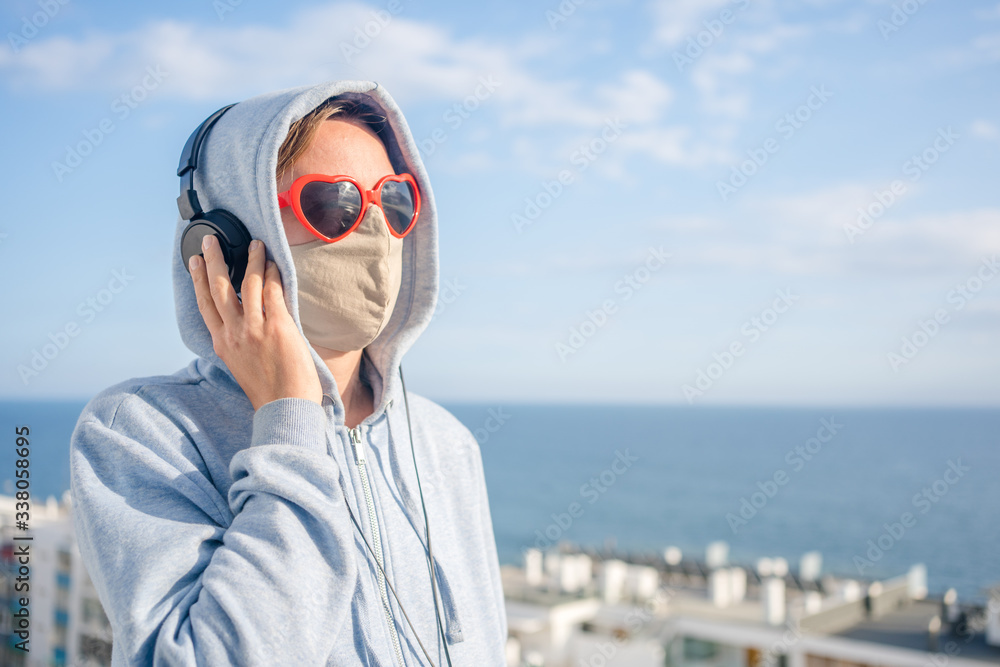 Female wearing protective mask and red love heart shape sunglasses using headphones listening music on outdoors background. Message of hope, having fun creative self-isolation. Closeup portrait person Female wearing protective mask and red love heart shape sunglasses using headphones listening music on outdoors background. Message of hope, having fun creative self-isolation. Closeup portrait person