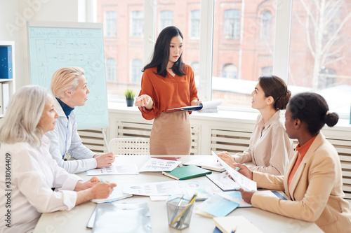 Portrait of successful Asian businesswoman presenting project plan to group of female colleagues while standing by whiteboard during meeting in conference room