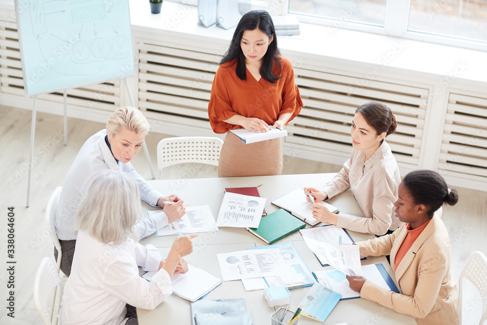 High angle view at successful Asian businesswoman presenting project plan to group of female colleagues while standing by whiteboard during meeting in conference room High angle view at successful Asian businesswoman presenting project plan to group of female colleagues while standing by whiteboard during meeting in conference room