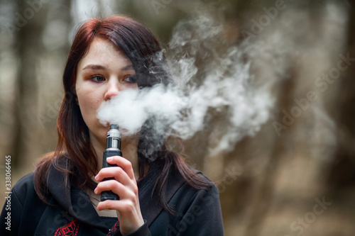 Vape teenager. Young pretty caucasian brunette girl smoking an electronic cigarette on the street in the spring. Deadly bad habit.