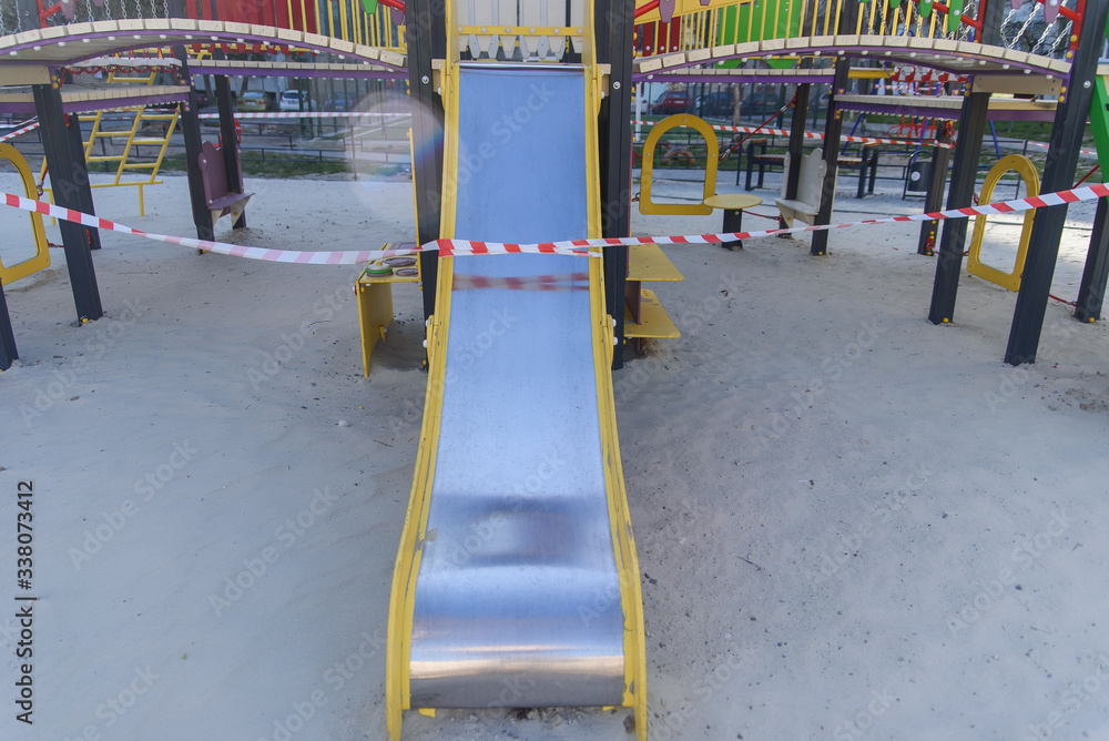 Fototapeta premium Coronavirus COVID-19 restriction. No people due to quarantine. Closed children's playground in the city. Empty park and playground. Stay at home compaign. Red warning tape on swings and slides