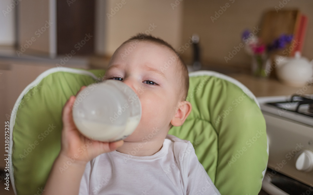 Baby boy sits in a feeding chair and drinks from a bottle. the boy in the kitchen. the baby eats the right food Baby boy sits in a feeding chair and drinks from a bottle. the boy in the kitchen. the baby eats the right food