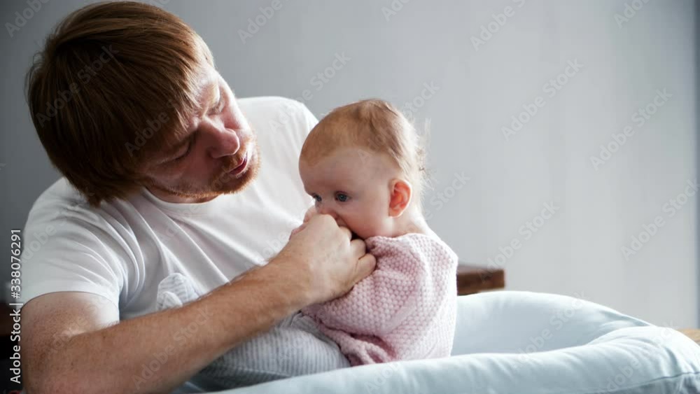 Careful dad talking to baby daughter sitting in her bedding. New father nursing little six month child at home. Medium shot. Baby care and parenthood concept Careful dad talking to baby daughter sitting in her bedding. New father nursing little six month child at home. Medium shot. Baby care and parenthood concept
