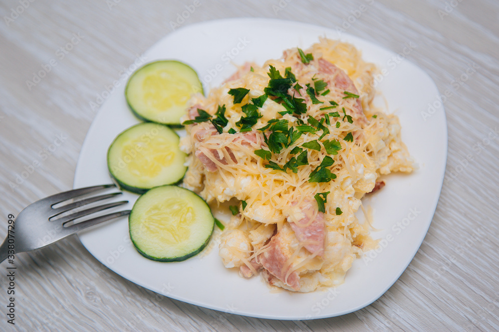 Omelet with sausage, herbs and cheese on a white plate. Dish with food on a light wooden table. Lunch fried eggs with bacon and chopped cucumber. Omelet with sausage, herbs and cheese on a white plate. Dish with food on a light wooden table. Lunch fried eggs with bacon and chopped cucumber.