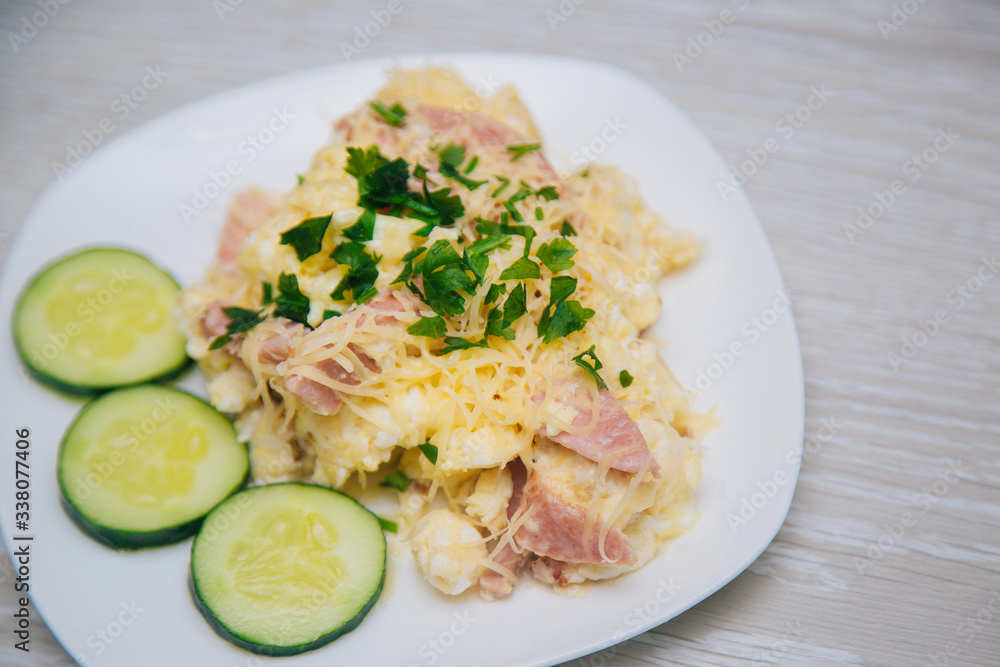 Omelet with sausage, herbs and cheese on a white plate. Dish with food on a light wooden table. Lunch fried eggs with bacon and chopped cucumber.