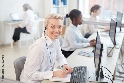 Portrait of female operator wearing headset and smiling at camera while sitting in row in call center or office interior, copy space