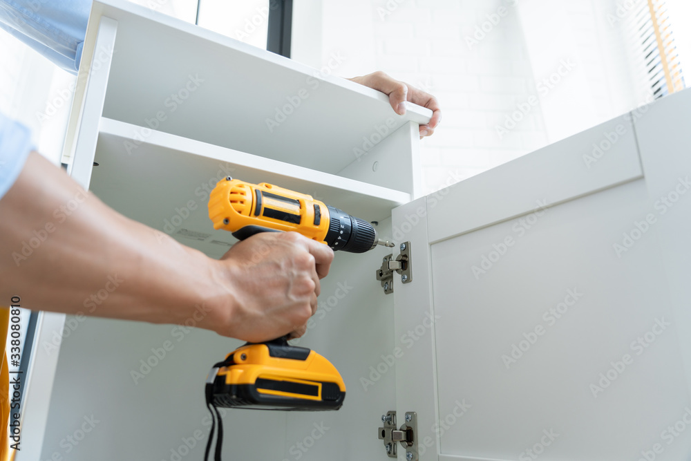 Close up portrait and details of caucasian male worker using electric screwdriver instrument in hand and repairing new wooden desk, home improvement concept.