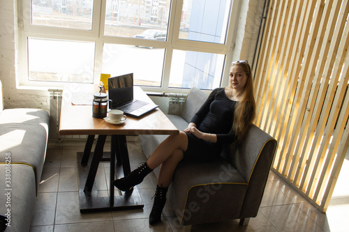 A beautiful girl in a black dress sits at a table by the window. Break work or computer training.