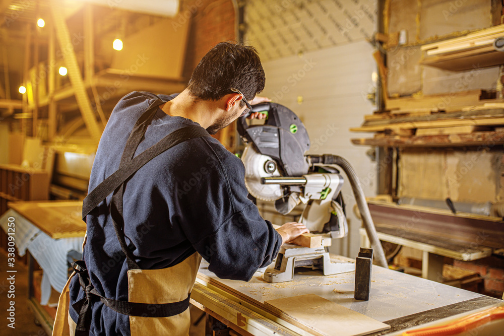 portrait of skilled young woodworker in safety gear using mitre or circular saw to cut piece of wood while working alone in his woodworking studio portrait of skilled young woodworker in safety gear using mitre or circular saw to cut piece of wood while working alone in his woodworking studio