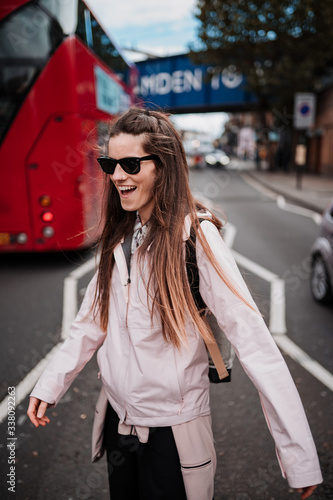 An attractive young woman with long brown hair, sunglasses and a pink coat smiles as she walks between two lanes of vehicles. In the background you can see a typical London bus.