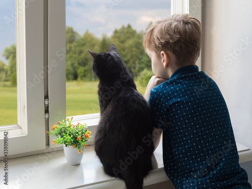 Caucasian boy and black cat look out the window. On the street flowering trees and good weather. Insulation concept in coronavirus and polynosis. Copyspace