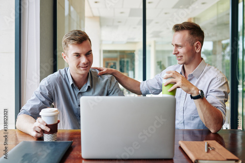 Smiling young businessman tapping colleague on shoulder and congratulating him with finishing big project
