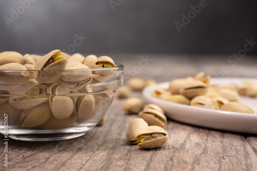 A closeup view of pistachio nuts in a small clear glass condiment bowl and on a white plate, in a lifestyle setting.