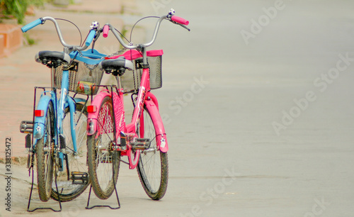 Two old colourful bicycles, one blue and one pink, parked on the left side of the grey empty street; male - female concept, love and romance