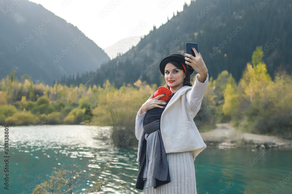 Fototapeta premium Beautiful young mother with her infant baby in sling make selfie. Mother is carrying her child and travel in autumn mountain lake. Active mother concept