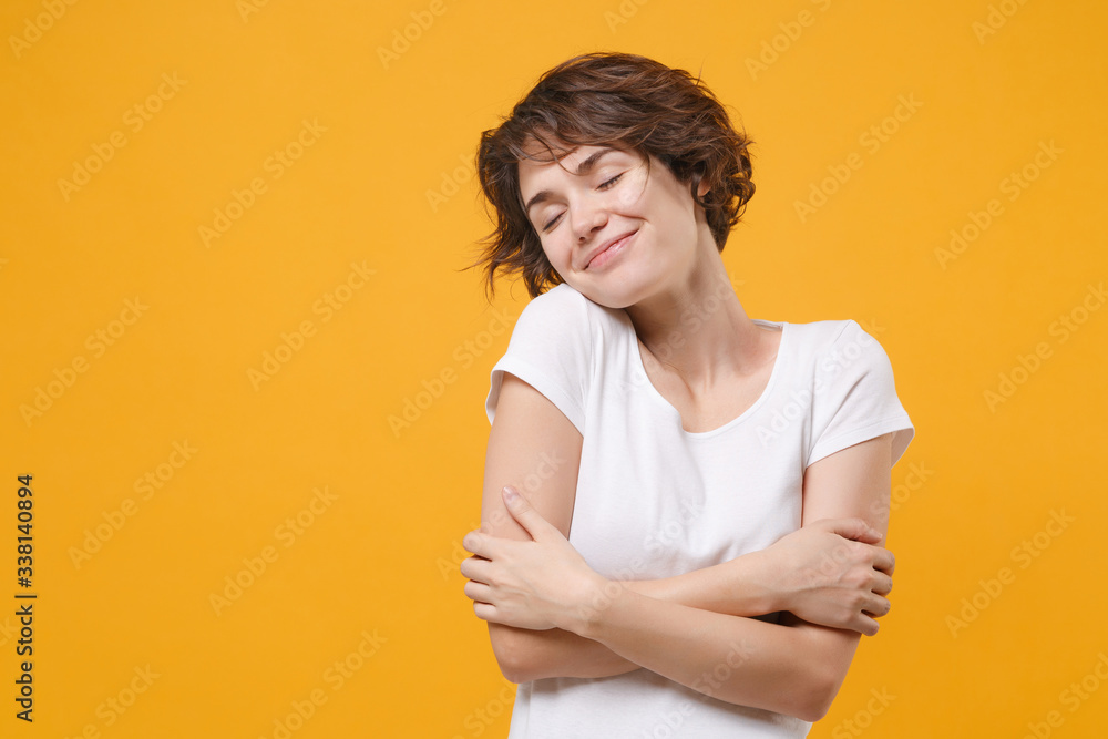 Obraz premium Smiling young brunette woman girl in white t-shirt posing isolated on yellow orange background. People lifestyle concept. Mock up copy space. Keeping eyes closed holding hands crossed hugging herself.