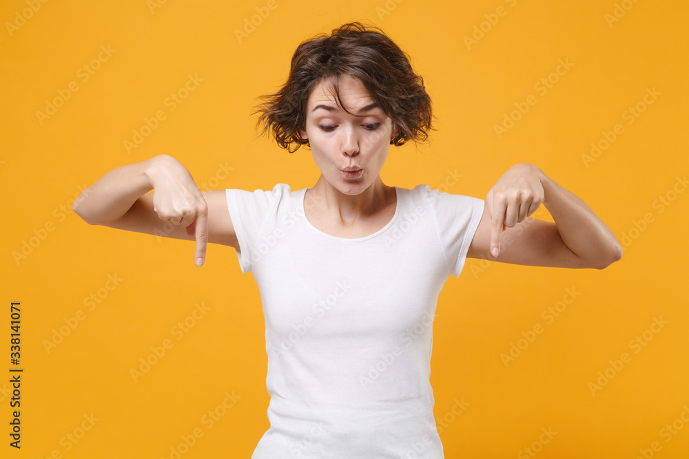Amazed young brunette woman in white t-shirt posing isolated on yellow orange wall background studio portrait. People sincere emotions lifestyle concept. Mock up copy space. Point index fingers down. Amazed young brunette woman in white t-shirt posing isolated on yellow orange wall background studio portrait. People sincere emotions lifestyle concept. Mock up copy space. Point index fingers down.