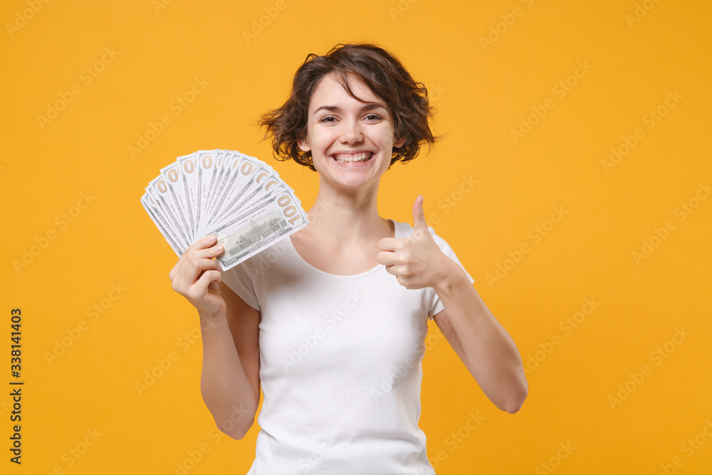 Smiling young brunette woman in white t-shirt posing isolated on yellow orange background. People lifestyle concept. Mock up copy space. Hold fan of cash money in dollar banknotes showing thumb up. Smiling young brunette woman in white t-shirt posing isolated on yellow orange background. People lifestyle concept. Mock up copy space. Hold fan of cash money in dollar banknotes showing thumb up.
