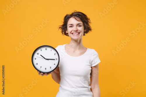 Smiling young brunette woman girl in white t-shirt posing isolated on yellow orange wall background studio portrait. People sincere emotions lifestyle concept. Mock up copy space. Hold in hand clock.