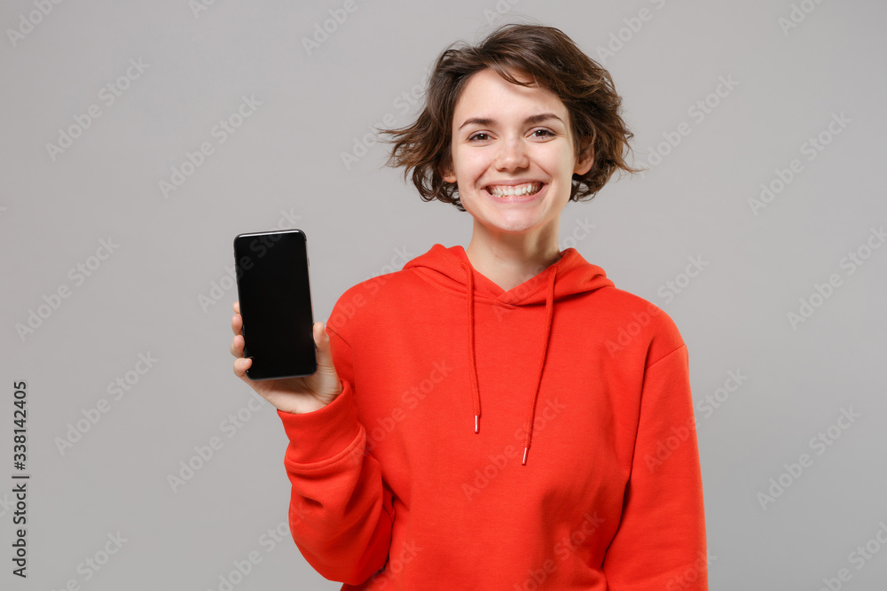 Smiling young brunette woman girl in casual red hoodie posing isolated on grey wall background studio portrait. People lifestyle concept. Mock up copy space. Hold mobile phone with blank empty screen. Smiling young brunette woman girl in casual red hoodie posing isolated on grey wall background studio portrait. People lifestyle concept. Mock up copy space. Hold mobile phone with blank empty screen.
