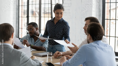 Young Indian businesswoman lead meeting with diverse colleagues share handouts at briefing, confident female biracial team leader head team discussion in office, consider paperwork with coworkers