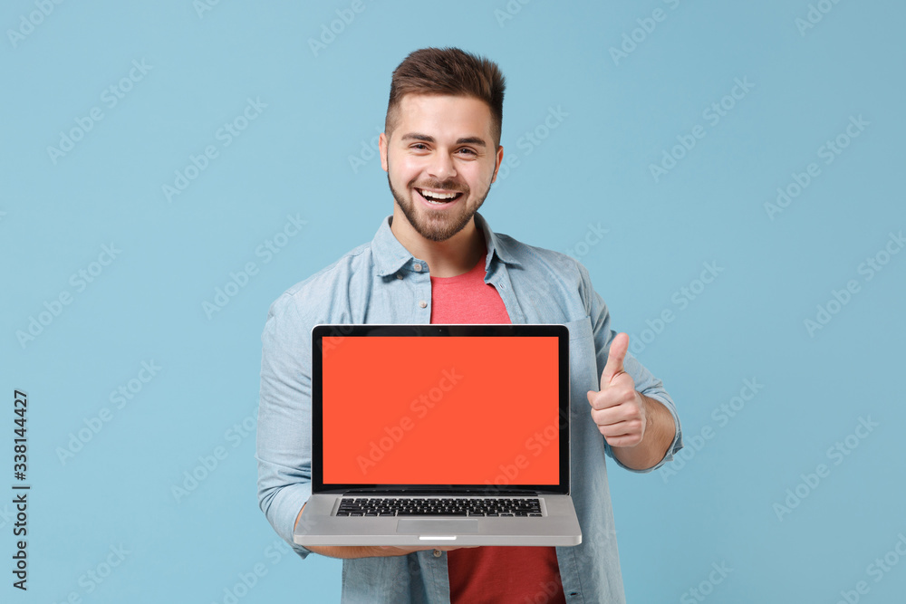Cheerful young bearded guy in casual shirt posing isolated on pastel blue background. People lifestyle concept. Mock up copy space. Hold laptop pc computer with blank empty screen, showing thumb up. Cheerful young bearded guy in casual shirt posing isolated on pastel blue background. People lifestyle concept. Mock up copy space. Hold laptop pc computer with blank empty screen, showing thumb up.