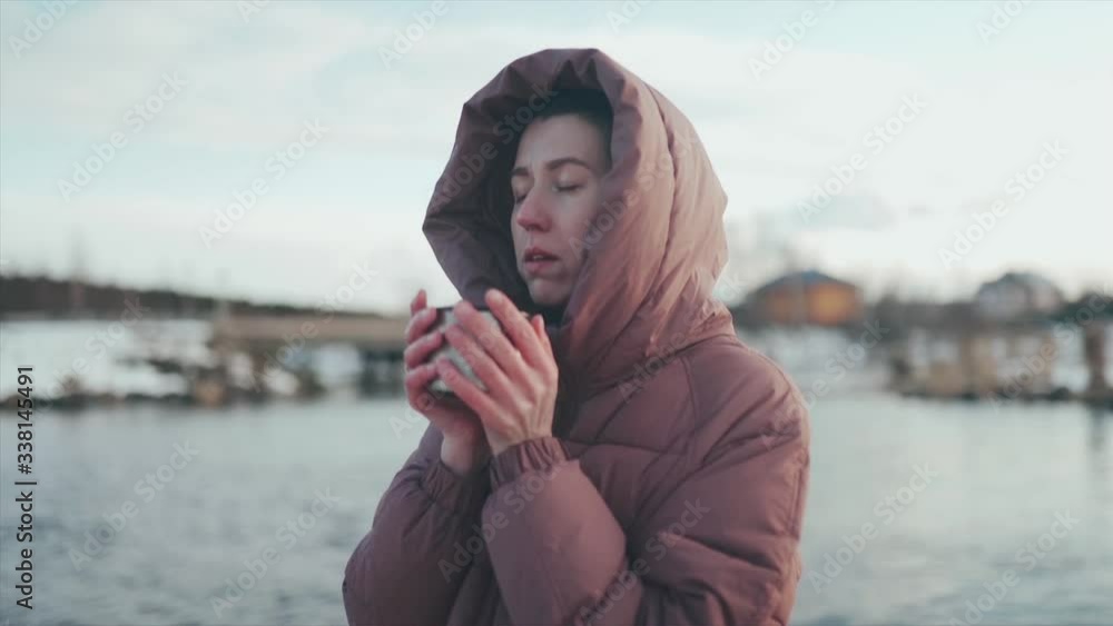 Close-up. A beautiful Caucasian woman drinks coffee standing on the Bank of a river in winter. She's wearing a dark jacket with a hood over her head. She takes a SIP of hot coffee and warms up. Close-up. A beautiful Caucasian woman drinks coffee standing on the Bank of a river in winter. She's wearing a dark jacket with a hood over her head. She takes a SIP of hot coffee and warms up.