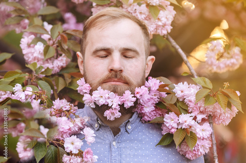 brutal bearded man in the flowering cherry trees. Handsome smiling bearded man outdoors. Spring day. Spring pink sakura blossom. Male spring fashion. Bearded stylish man. Happy guy in pink flowers