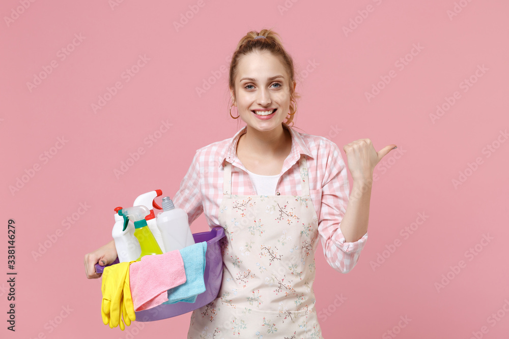 Smiling young woman housewife in apron hold basin with detergent bottles washing cleansers while doing housework isolated on pink background studio portrait. Housekeeping concept. Point thumb aside. Smiling young woman housewife in apron hold basin with detergent bottles washing cleansers while doing housework isolated on pink background studio portrait. Housekeeping concept. Point thumb aside.