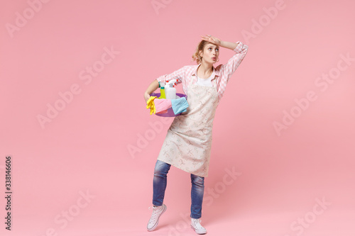 Tired young woman housewife in apron hold basin with detergent bottles washing cleansers while doing housework isolated on pink background studio portrait. Housekeeping concept. Put hand on head.