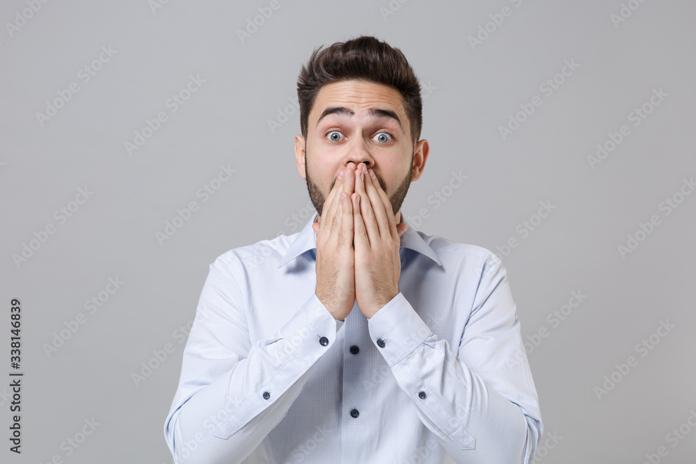 Shocked young unshaven business man in light shirt posing isolated on grey wall background studio portrait. Achievement career wealth business concept. Mock up copy space. Covering mouth with hands. Shocked young unshaven business man in light shirt posing isolated on grey wall background studio portrait. Achievement career wealth business concept. Mock up copy space. Covering mouth with hands.