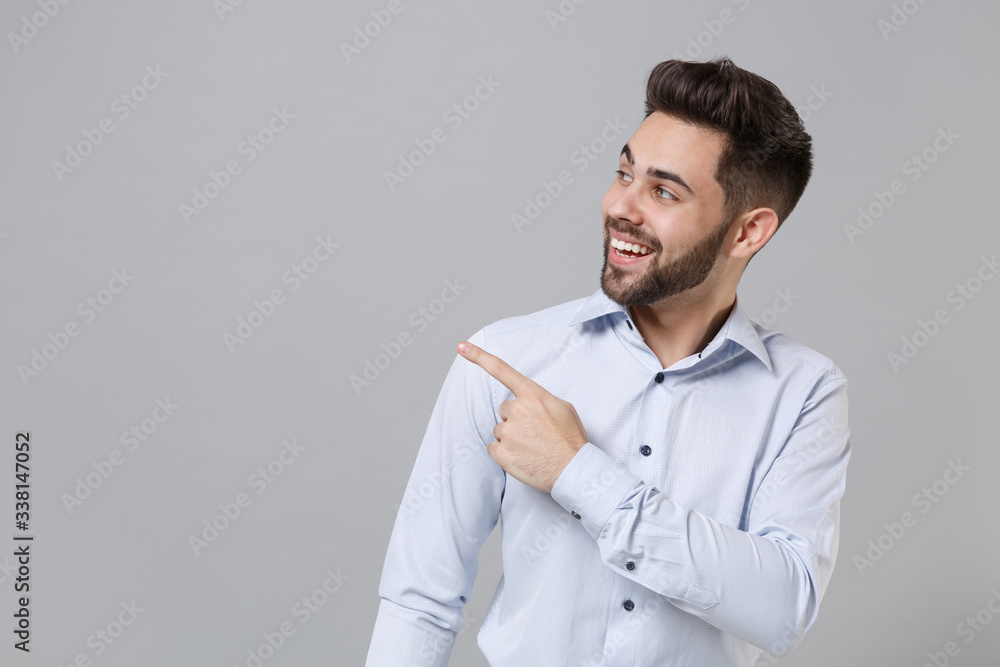 Smiling young unshaven business man in light shirt posing isolated on grey background studio portrait. Achievement career wealth business concept. Mock up copy space. Pointing index finger aside up. Smiling young unshaven business man in light shirt posing isolated on grey background studio portrait. Achievement career wealth business concept. Mock up copy space. Pointing index finger aside up.