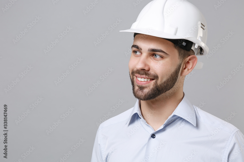 Smiling young unshaven business man in light shirt protective construction helmet posing isolated on grey background. Achievement career wealth business concept. Mock up copy space. Looking aside. Smiling young unshaven business man in light shirt protective construction helmet posing isolated on grey background. Achievement career wealth business concept. Mock up copy space. Looking aside.