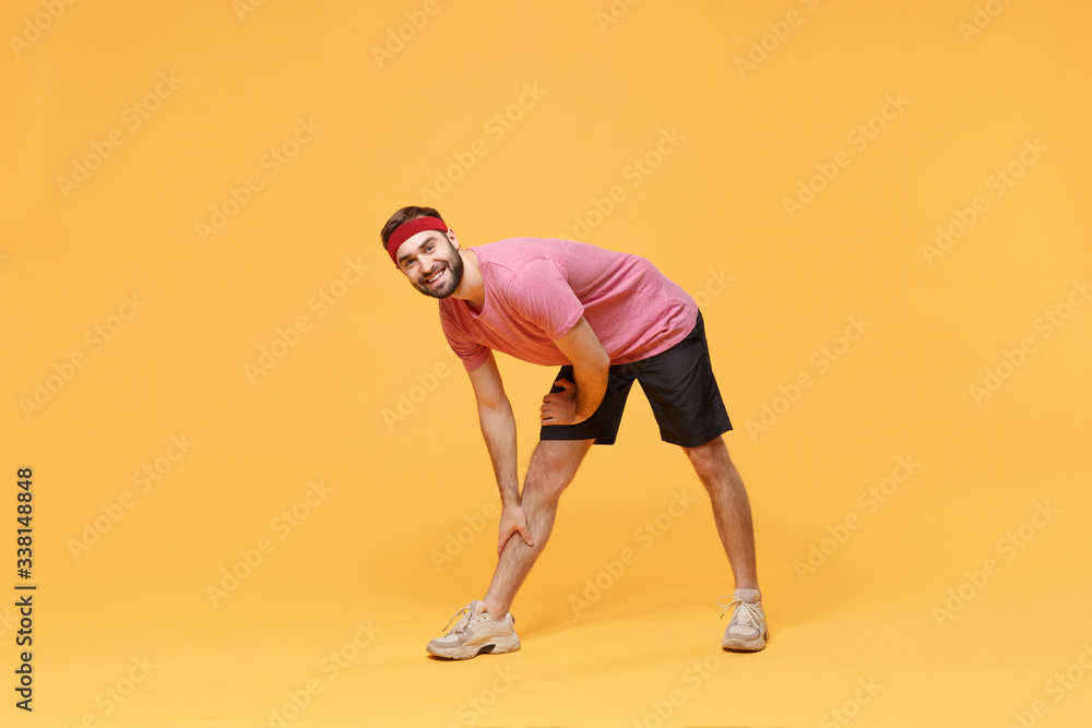 Smiling young bearded fitness sporty guy sportsman in headband t-shirt in home gym isolated on yellow background. Workout sport motivation concept. Doing warm up, stretching exercising, leaning aside. Smiling young bearded fitness sporty guy sportsman in headband t-shirt in home gym isolated on yellow background. Workout sport motivation concept. Doing warm up, stretching exercising, leaning aside.