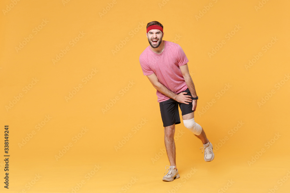 Fototapeta premium Ingured displeased young bearded fitness guy 20s sportsman in headband t-shirt in home gym isolated on yellow wall background. Workout sport motivation concept. Touch leg with elastic bandage on knee.