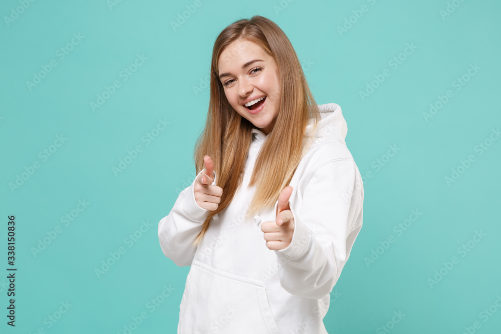 Fototapeta premium Cheerful young woman girl in casual white hoodie posing isolated on blue turquoise background studio portrait. People emotions lifestyle concept. Mock up copy space. Pointing index fingers on camera.