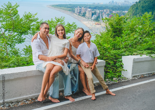 Cheerful, happy family, father, mother, son and daughter enjoying walk play outdoor near sea. Family in white clothes barefoot on walkway. Green forest background.