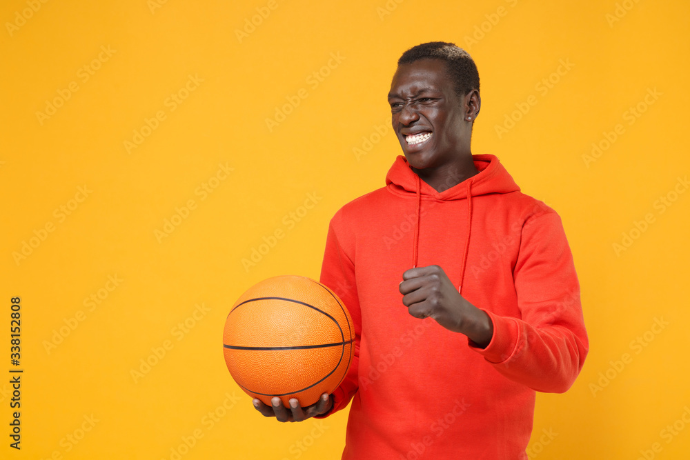 Irritated young african american man guy basketball player in red streetwear hoodie posing isolated on yellow background. Sport leisure lifestyle concept. Playing basketball hold ball clenching fist. Irritated young african american man guy basketball player in red streetwear hoodie posing isolated on yellow background. Sport leisure lifestyle concept. Playing basketball hold ball clenching fist.