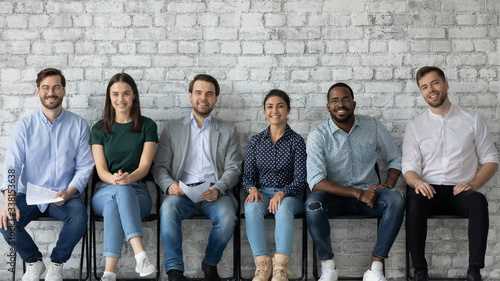 Portrait of smiling diverse multiracial young people sit in row on chairs wait for office interview, happy motivated multiethnic job candidates hired for vacant work position, employment concept