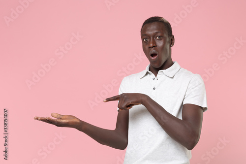 Shocked young african american man guy in white polo shirt posing isolated on pastel pink background studio portrait. People lifestyle concept. Mock up copy space. Pointing index fingers hand aside.